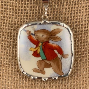 May include: A silver-toned pendant necklace featuring a square pendant with a cartoon rabbit illustration. The rabbit is wearing a red jacket, green shirt, and yellow tie. The pendant is framed in silver and hangs from a silver chain.