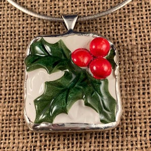 May include: A square pendant featuring a glossy white background with a raised, hand-painted design of dark green holly leaves and three bright red berries. The piece is encased in a silver-toned metal border and hangs from a silver wire necklace.