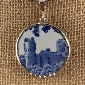 May include: A round pendant necklace with a silver-colored bezel. The pendant features a blue and white design of a building and foliage. The necklace is on a silver-colored chain.