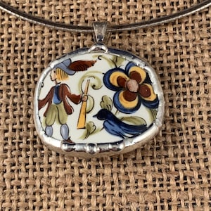 May include: A pendant necklace featuring a ceramic shard set in a silver-toned metal frame. The shard displays a hand-painted illustration of a person in a hat holding a staff, a large blue and yellow flower, and a small blue bird.