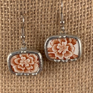 May include: A pair of handmade earrings featuring rectangular ceramic tiles with a floral design in shades of orange and white. The tiles are encased in a silver-colored metal frame, and the earrings have silver-colored fish hook ear wires.