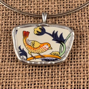 May include: A pendant featuring a piece of cream-colored ceramic with a graphic illustration of a yellow and orange bird perched on a branch, surrounded by dark blue floral elements, all encased in a silver-toned metal frame on a chain.