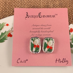 May include: A pair of clip-on earrings featuring a vintage china design with a holly and berry motif. The earrings are silver-toned and have a rectangular shape. The card reads "Antique Chinawear™ Antique china from around the world beautifully handcrafted for today's woman. Clip Holly"