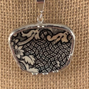 May include: A silver pendant necklace featuring a rectangular pendant with a black and white floral pattern. The pendant is framed in silver and hangs from a silver chain. The necklace is displayed against a neutral background.