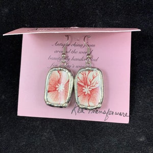 May include: A pair of earrings made from antique china with a floral design in red and white. The earrings are handcrafted and have silver-toned metal settings. The card reads "Antique china from around the world beautifully handcrafted for today's woman" and "Red Transferware".