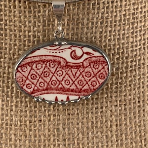 May include: Oval pendant necklace featuring a red and white patterned ceramic design. The pendant is encased in a silver-toned metal frame and hangs from a silver chain. The ceramic design has a repeating diamond pattern.