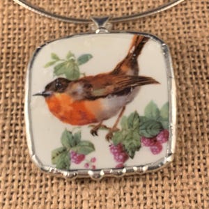May include: A square pendant necklace featuring a ceramic tile with a detailed illustration of a robin perched on a branch with green leaves and small pink berries. The tile is encased in a silver-toned metal frame and hangs from a thin silver wire.