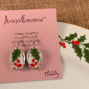 May include: A pair of oval-shaped dangle earrings featuring a white background with green holly leaves and red berries, encased in a silver-toned metal frame. The earrings are attached to a pink display card labeled Antique Chinawear and Holly.