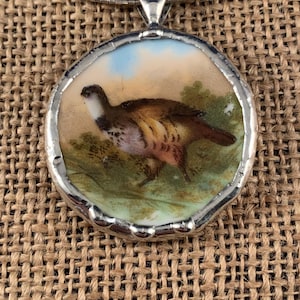 May include: A round pendant necklace featuring a hand-painted illustration of a brown and white game bird in a grassy landscape. The circular ceramic piece is encased in a silver-toned metal frame with a smooth, polished finish and a bail.