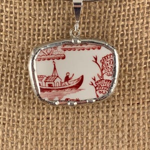 May include: A silver-toned necklace with a rectangular pendant. The pendant features a red and white design of a boat on a river, with trees and buildings. The pendant is framed in silver.