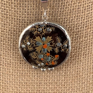 May include: A round pendant with a silver-colored frame and bail. The pendant features a dark background with a floral design in gold, blue, and orange. The pendant hangs from a silver-colored chain.
