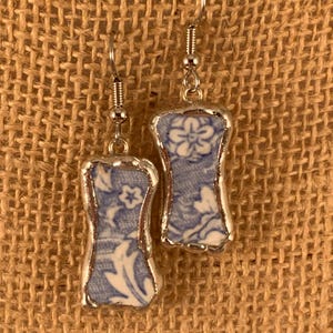 May include: A pair of dangle earrings featuring rectangular ceramic shards with rounded edges, decorated in a blue and white floral pattern. The ceramic pieces are encased in a silver-toned metal border and hang from silver-toned metal hooks.