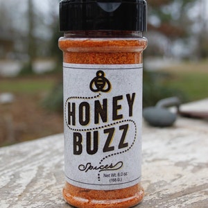 May include: A clear spice jar with a black lid, filled with orange-colored spices. The label reads "HONEY BUZZ Spices" with a bee logo. Net weight is 6.0 oz (168 g). The jar is set on a weathered wooden surface.