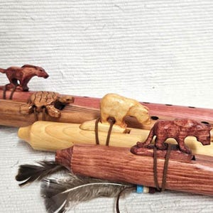 May include: A collection of wooden Native American flutes, each adorned with a carved animal figure. The flutes vary in color, from light wood to reddish-brown, and feature carvings of a bear, turtle, and other animals. Feathers are attached to some flutes.