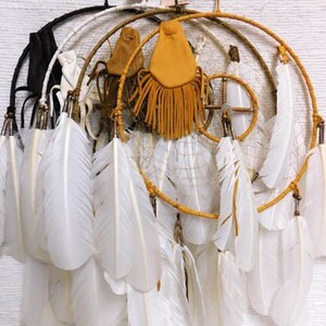 May include: A set of three dreamcatchers with white feathers hanging from them. The dreamcatchers are made of wood and have a brown, tan, and white color scheme. The dreamcatchers are hanging from a white wall.