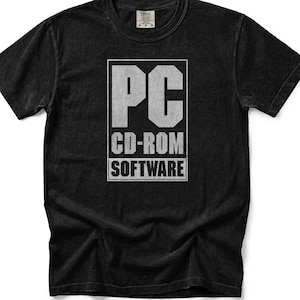 Op de afbeelding: Een zwart T-shirt met korte mouwen en een witte rechthoekige afbeelding op de borst. De afbeelding bevat de tekst PC in grote letters, gevolgd door CD-ROM en SOFTWARE in kleinere gestapelde tekst, alles binnen een dunne witte rand.