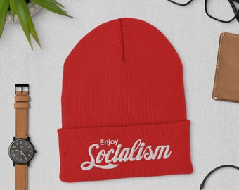 May include: A vibrant red beanie hat featuring the embroidered phrase "Enjoy Socialism" in white script. The hat's brim is folded, and it's made from a soft, knitted fabric.