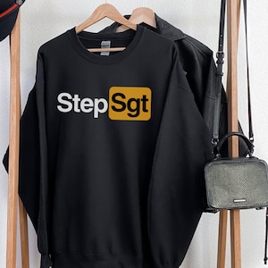 Puede incluir: Sudadera negra con el texto "Step Sgt" en letras blancas dentro de un cuadrado dorado. La sudadera est&aacute; colgada en un perchero de madera con un sombrero negro y un bolso negro.