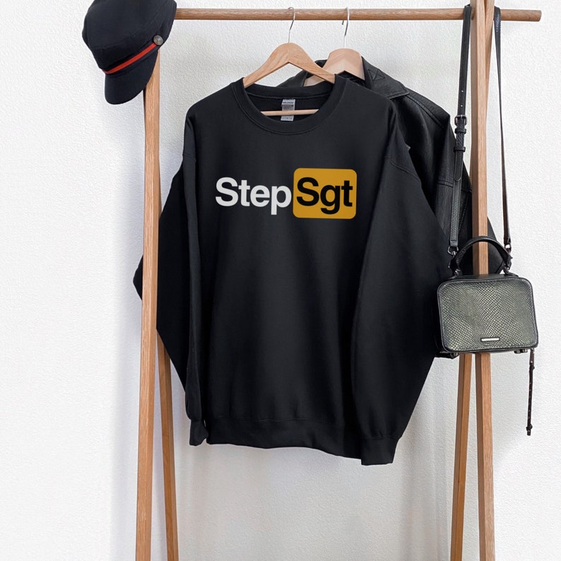 Puede incluir: Sudadera negra con el texto "Step Sgt" en letras blancas dentro de un cuadrado dorado. La sudadera est&aacute; colgada en un perchero de madera con un sombrero negro y un bolso negro.