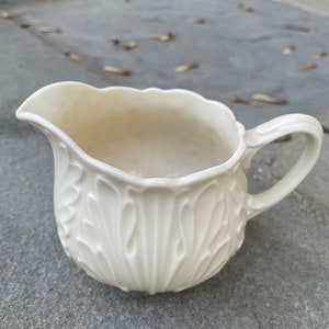 May include: A white ceramic creamer with an ornate, embossed design. The creamer has a rounded body and a curved handle.