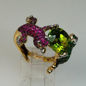 May include: A gold ring with two lizards, one with pink and white stones and the other with green and white stones. The lizards are holding a large green gemstone.