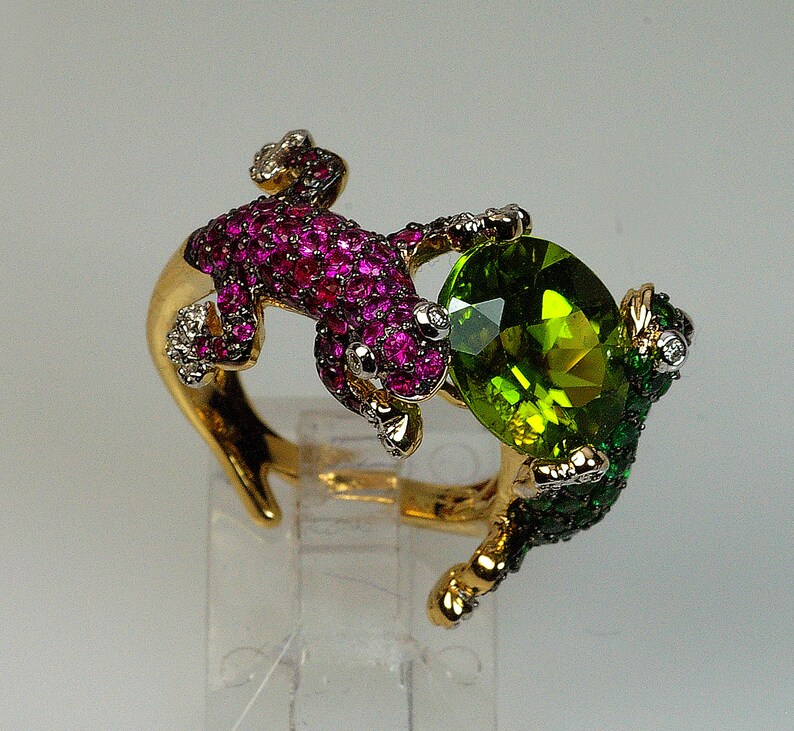 May include: A gold ring with two lizards, one with pink and white stones and the other with green and white stones. The lizards are holding a large green gemstone.