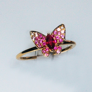 May include: A gold ring with a pink and white butterfly design. The butterfly has a red ruby in the center and is surrounded by smaller pink and white stones.
