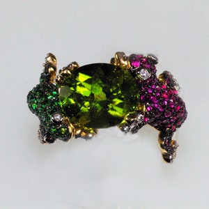 May include: A ring with a large green gemstone surrounded by two frogs, one with green and black accents and the other with pink and red accents.
