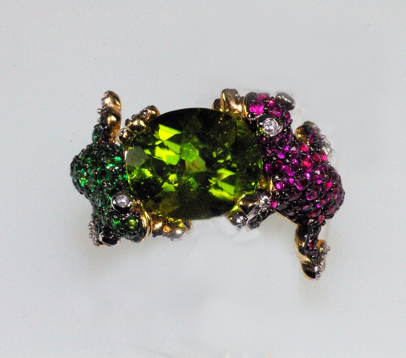 May include: A ring with a large green gemstone surrounded by two frogs, one with green and black accents and the other with pink and red accents.