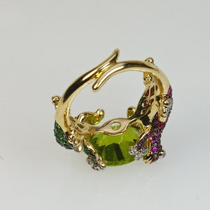 May include: Gold ring with a large green gemstone surrounded by smaller red and green gemstones. The ring is set in a gold band with a delicate design.