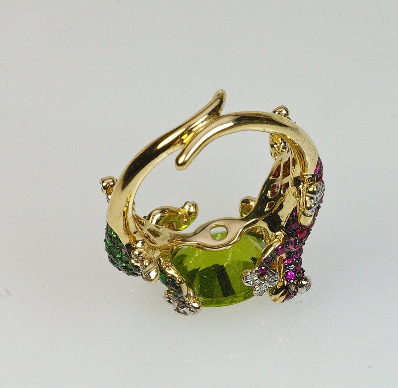 May include: Gold ring with a large green gemstone surrounded by smaller red and green gemstones. The ring is set in a gold band with a delicate design.