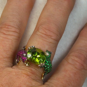 May include: A gold ring with two frog figures on either side of a large green peridot gemstone. The frogs are set with smaller green and red gemstones.