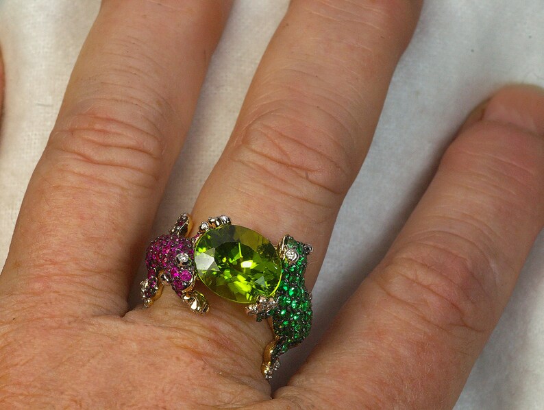 May include: A gold ring with two frog figures on either side of a large green peridot gemstone. The frogs are set with smaller green and red gemstones.