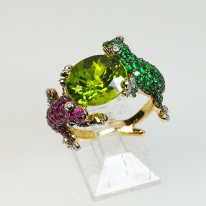 May include: A gold ring with two frog-shaped settings, one with green and one with pink gemstones. The frogs are holding a large, round, green peridot gemstone in the center.