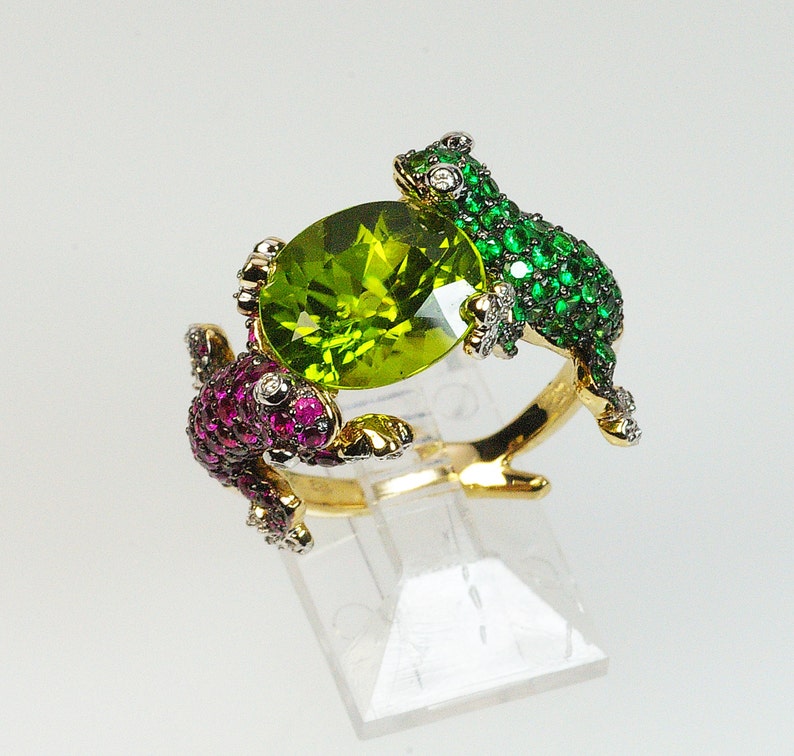 May include: A gold ring with two frog-shaped settings, one with green and one with pink gemstones. The frogs are holding a large, round, green peridot gemstone in the center.