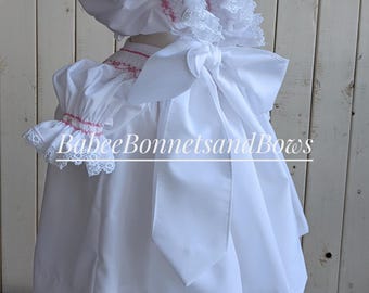 May include: White baby dress with pink embroidery and a white bonnet with pink embroidery and lace trim. The dress has a large white bow in the back. The dress is on a wooden mannequin. "BabeeBonnetsandBows" is written on the dress.