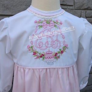 May include: White long-sleeved dress with a pink embroidered monogram and a birthday cake design. The dress has a pink skirt and a white bodice with a lace trim. The text "BabeeBonnetsandBows" is embroidered on the back.