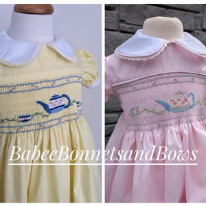 May include: Two smocked dresses for a baby or toddler, one yellow and one pink. Both dresses feature a white Peter Pan collar and a teacup and teapot embroidery design. The dresses are made by BabeeeBonnetsandBows.