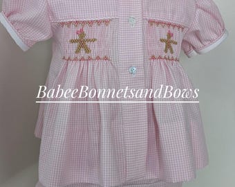 May include: A pink and white gingham short-sleeve shirt and bloomer set featuring a Peter Pan collar with lace trim, three front buttons, and smocking on the chest with embroidered gingerbread figures. The text BabeeBonnetsandBows is visible.