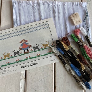 May include: A cross-stitch kit featuring a girl and kittens. The kit includes a white fabric, floss in various colors, and a pattern with the title "Katie's Kittens".
