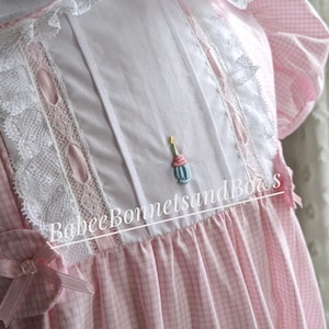 May include: A pink and white gingham dress with a white collar and lace trim. The dress features a decorative embroidered cupcake and the text "Babee Bonnets and Bows". The dress has short sleeves and a bow detail.