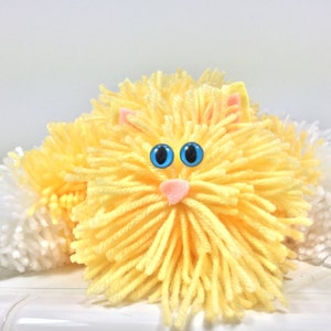 May include: A yellow yarn cat with white yarn tail pom-poms and blue eyes.