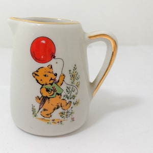May include: A white ceramic creamer with a gold rim and handle. It features a cartoon illustration of a bear holding a red balloon.