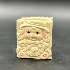May include: A small, rectangular, off-white ceramic container with a relief design of a cat. The cat has pink accents on its eyes, ears, and paws. The container has a scalloped top edge.
