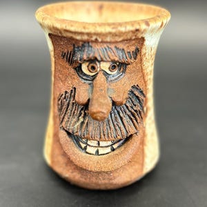 Könnte beinhalten: Eine handgefertigte Keramiktasse mit einem geschnitzten Gesichtsdesign. Die Tasse zeigt ein lächelndes Gesicht mit Schnurrbart, Augenbrauen und Augen. Die Tasse ist braun mit beigen Akzenten und hat eine zylindrische Form.