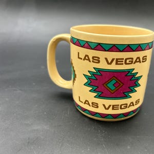 Może przedstawiać: Kubek ceramiczny w kolorze beżowym z turkusowym i różowym wzorem geometrycznym i nadrukiem "LAS VEGAS" dwa razy.