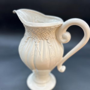 May include: A white ceramic pitcher with an ornate design. The pitcher has a wide base and a tall, slender body. The handle is curved and has a decorative scroll at the top.