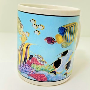 May include: A white ceramic mug with a blue background and colorful fish and coral illustrations. The mug features a variety of fish, including a yellow and blue angelfish, a purple and red fish, and a yellow and black fish. The mug also features a coral reef with red, orange, and yellow coral.