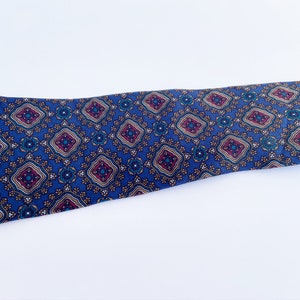 May include: A blue necktie with a repeating geometric pattern. The pattern features diamond shapes in burgundy, teal, and gold. The tie is made of a smooth fabric and is displayed on a white background.