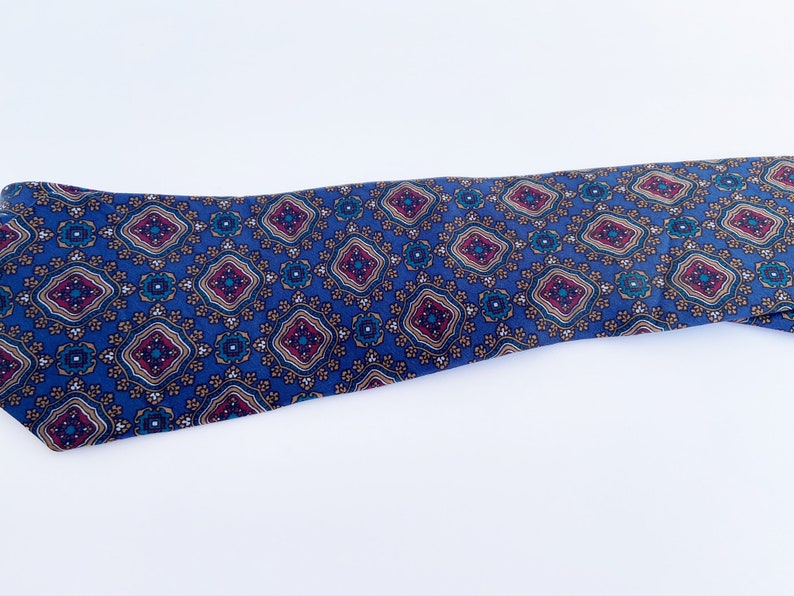 May include: A blue necktie with a repeating geometric pattern. The pattern features diamond shapes in burgundy, teal, and gold. The tie is made of a smooth fabric and is displayed on a white background.